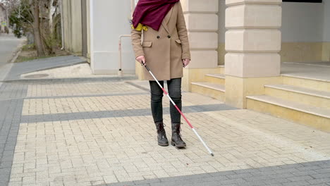 mid view of a blind woman in brown coat and scarf walking with a walking stick in the street