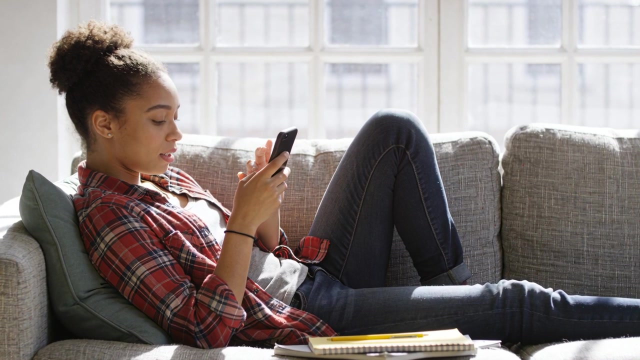 Une femme en flanelle rouge regardant un smartphone, faisant défiler et tapotant sur un canapé près de la fenêtre, avec un carnet.