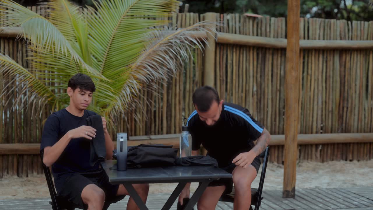 Two men packing or organizing items at an outdoor table in a tropical setting