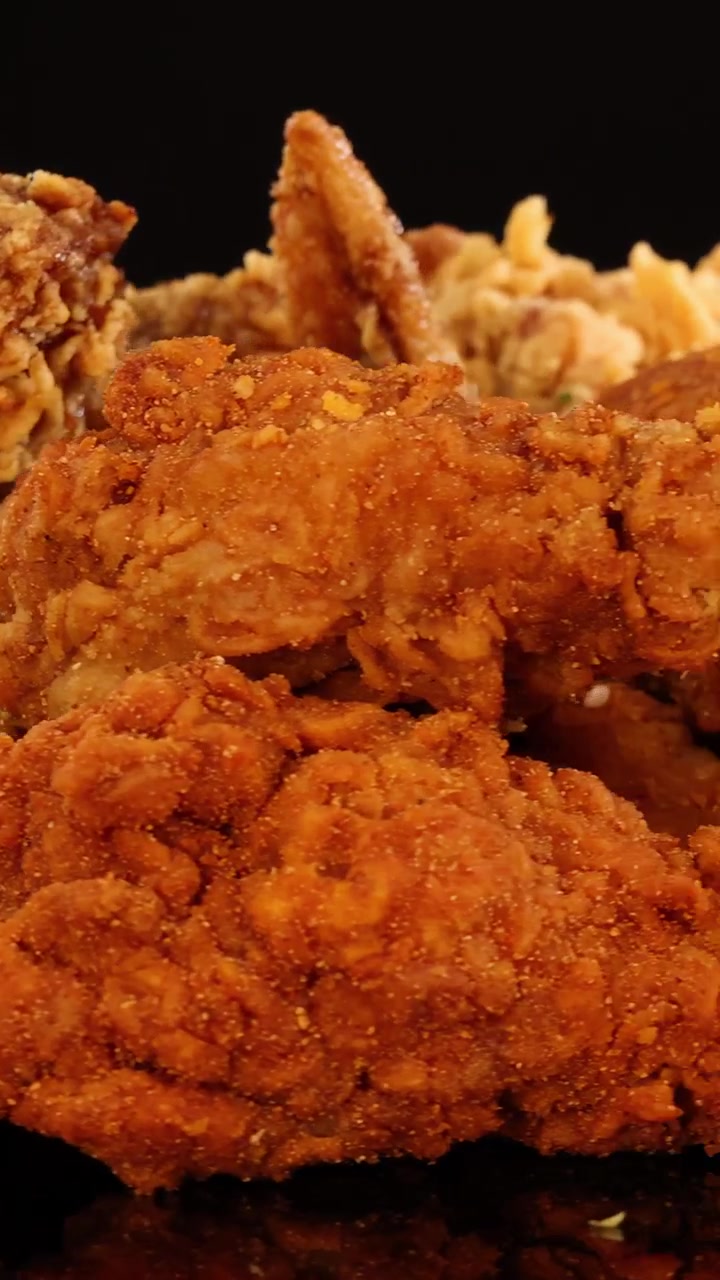 Alitas de pollo fritas doradas, muslitos y nuggets giran sobre una superficie negra brillante bajo iluminación de estudio, resaltando texturas crujientes y detalles apetitosos.