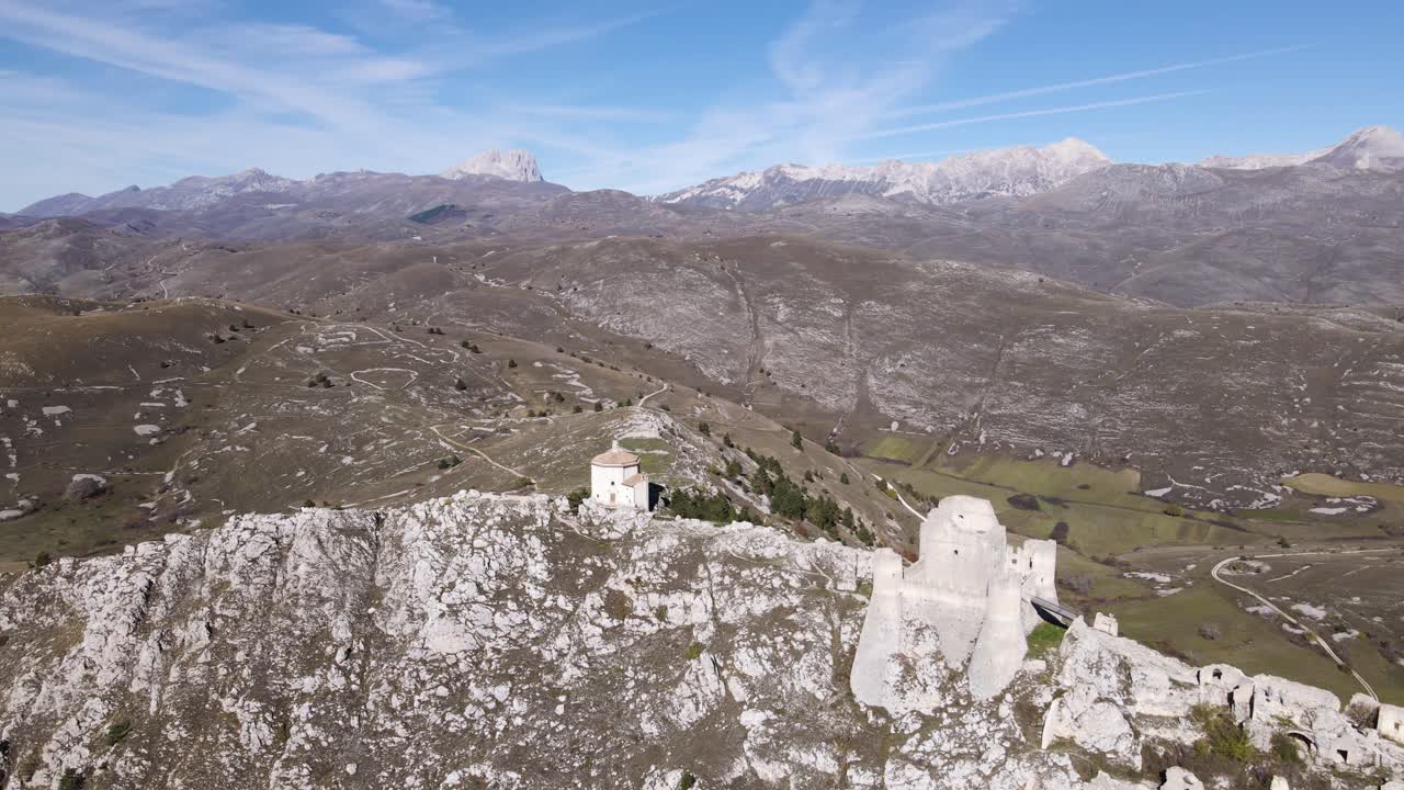 Veduta aerea della fortezza medievale di Rocca Calascio arroccata su una cresta di calcare bianco con ampie valli montane, paesaggio rurale e le cime del Gran Sasso sullo sfondo.