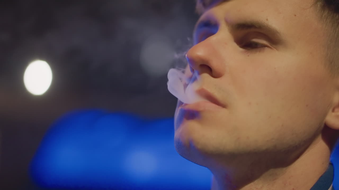 네온 불빛이 켜진 다리 근처에서 전자 담배를 피우는 백인 운전자, 손가락 사이에 들고 있는 휴대용 전자 담배, 교통 체증을 향한 강렬한 시선, 연기 고리가 보이는 중간 클로즈업, 야간 근무 중 길가에서 대기 중, 도시의 불빛