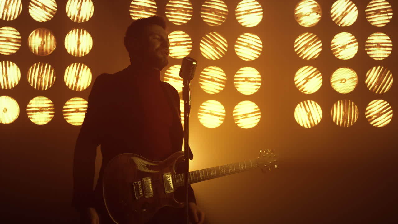 Gitaris bernyanyi dengan mikrofon, menikmati proses di bawah sorotan lampu. Pria menampilkan lagu.