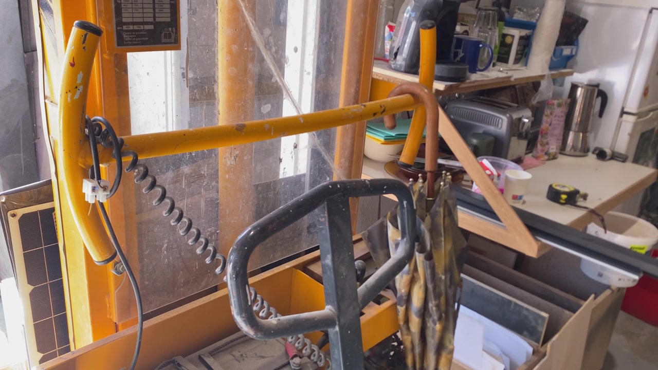 Yellow hand pallet truck with a coiled cable and a hanging brown umbrella, standing in a messy workshop or garage space