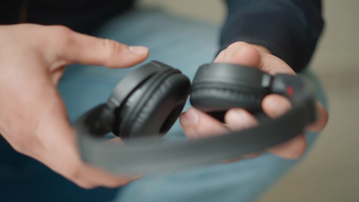 Primo piano di mani che esaminano cuffie over-ear nere, ruotando e ispezionando il dispositivo. La persona indossa jeans azzurri in un ambiente interno casual con illuminazione soffusa, concentrandosi sui dettagli della texture.