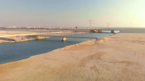 Pedestrian-bridge-over-blue-canal-in-barren-desert-environment,-aerial-view,-UAE