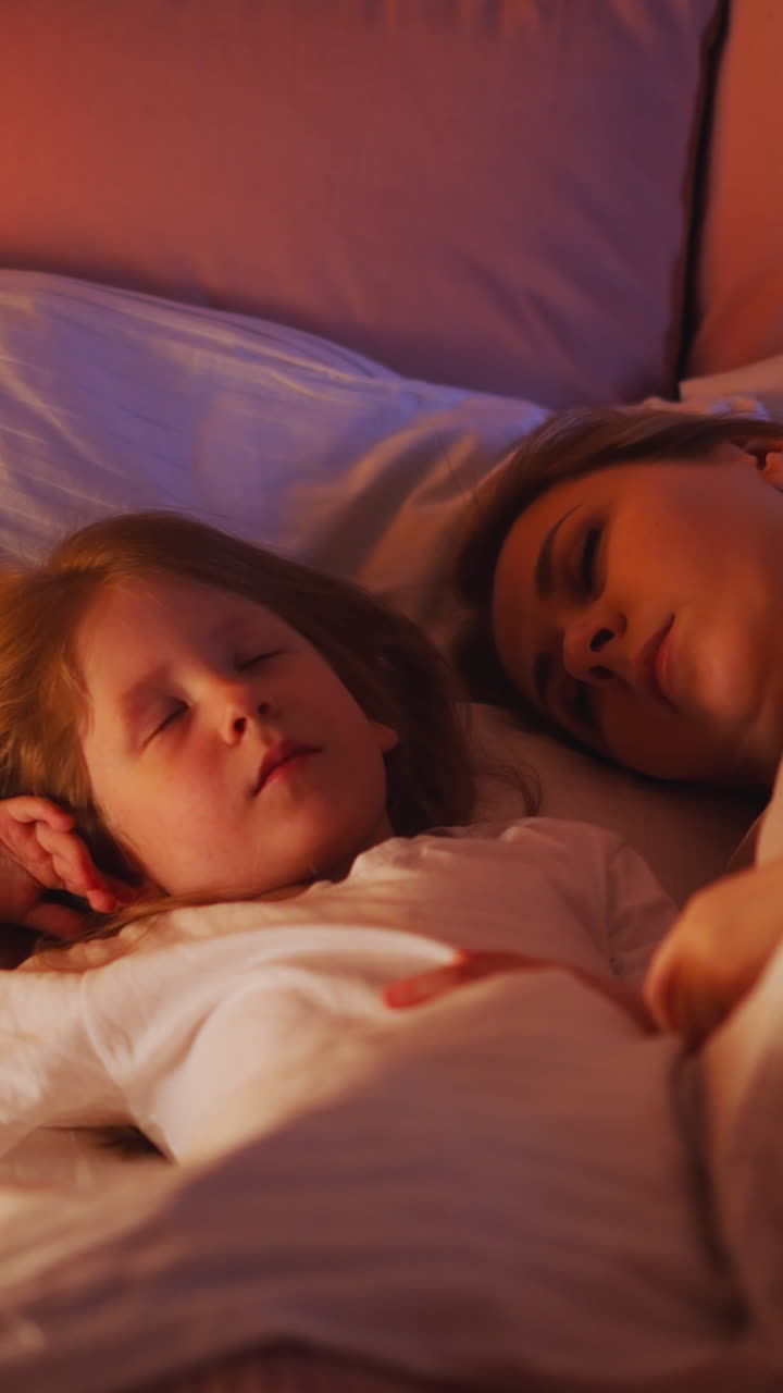 Pretty little girl and tired mommy sleep together in wide bed with soft pillows and warm comforter at warm light of glowing night lamp in bedroom slow motion