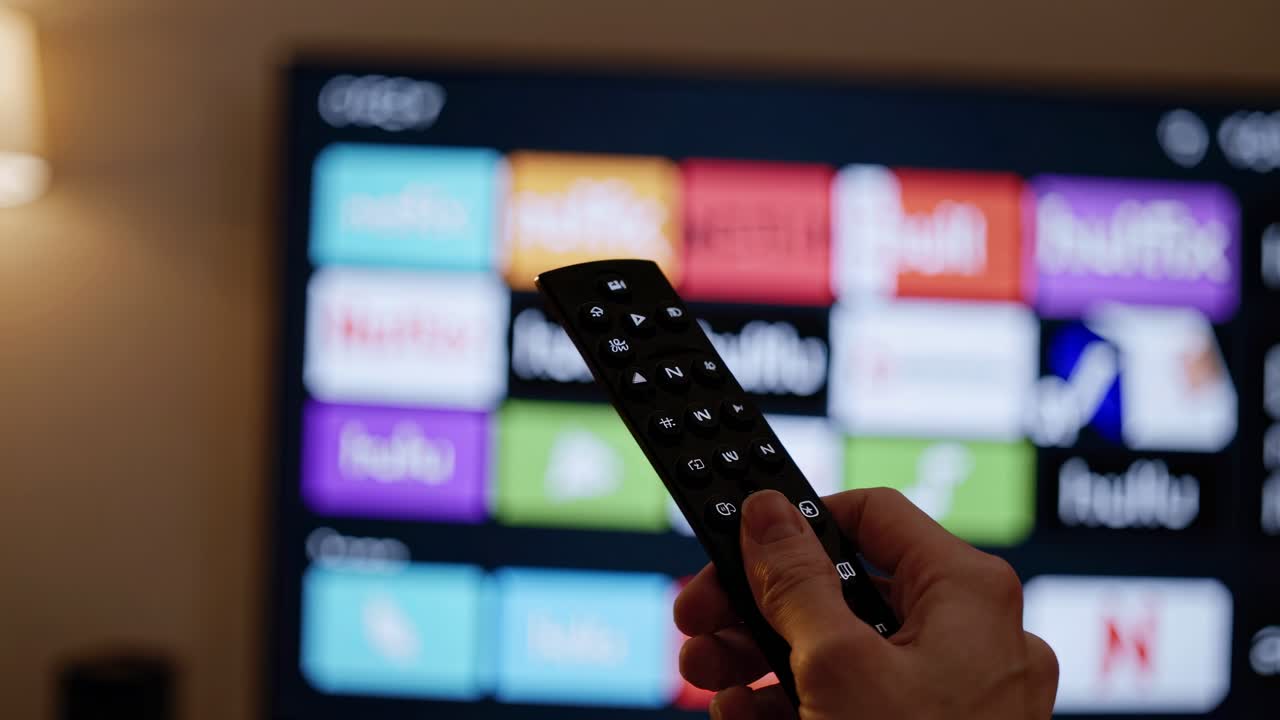 Primo piano di una mano che tiene un telecomando, puntato verso uno schermo TV che mostra vari video in streaming.