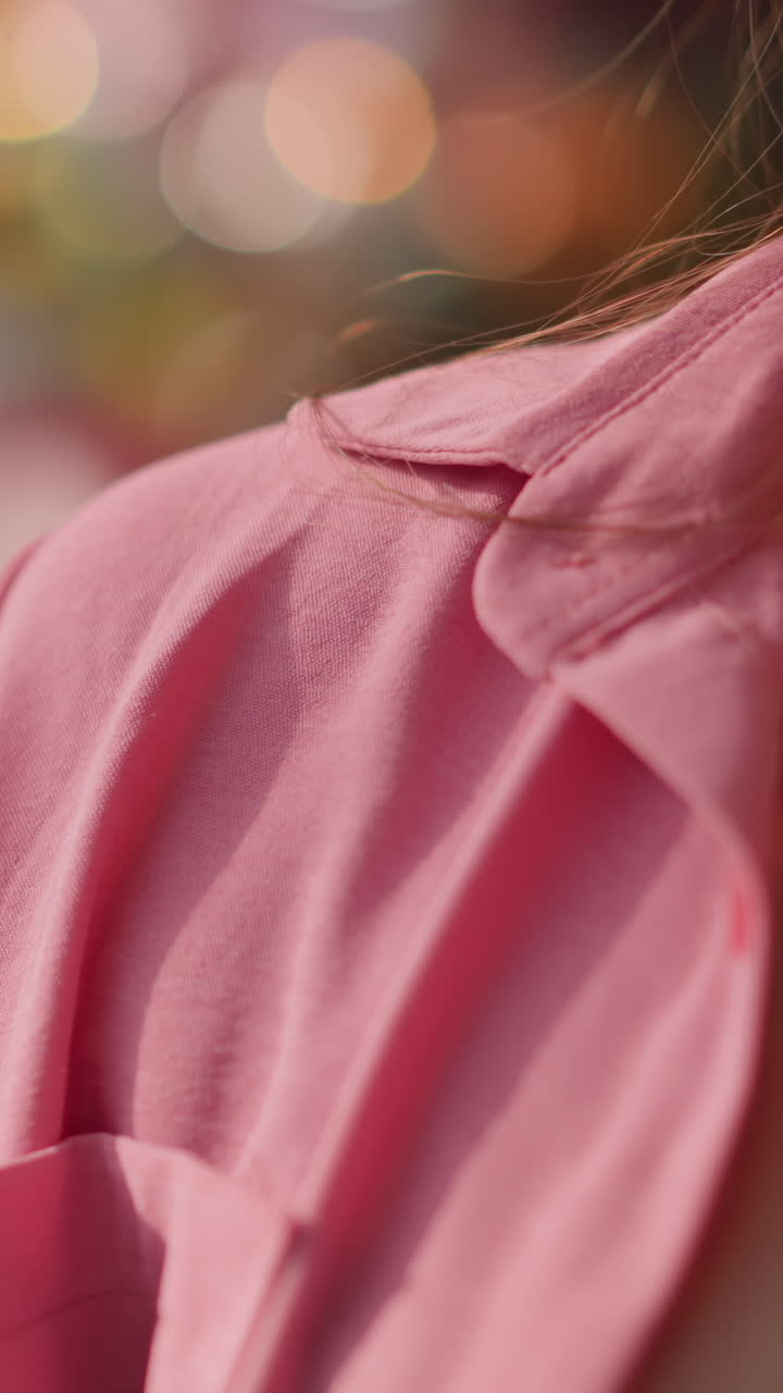 Lady wearing unbuttoned pink dress places her hand with a shopping bag behind her, the background features a blurred view of glowing lights, creating a soft and radiant effect around her silhouette