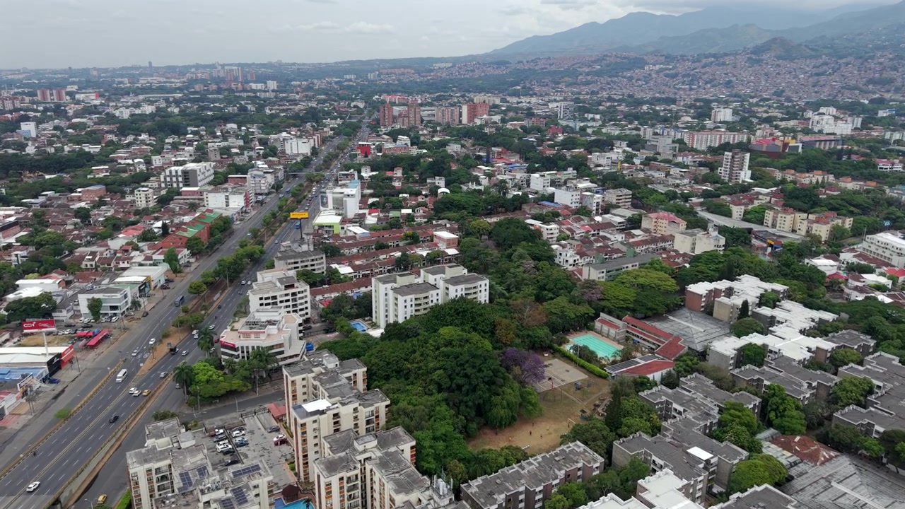 Dynamic drone video captures Cali’s Autopista Sur, showcasing traffic flow, residential buildings, urban greenery, and Andean mountains under cloudy daylight.