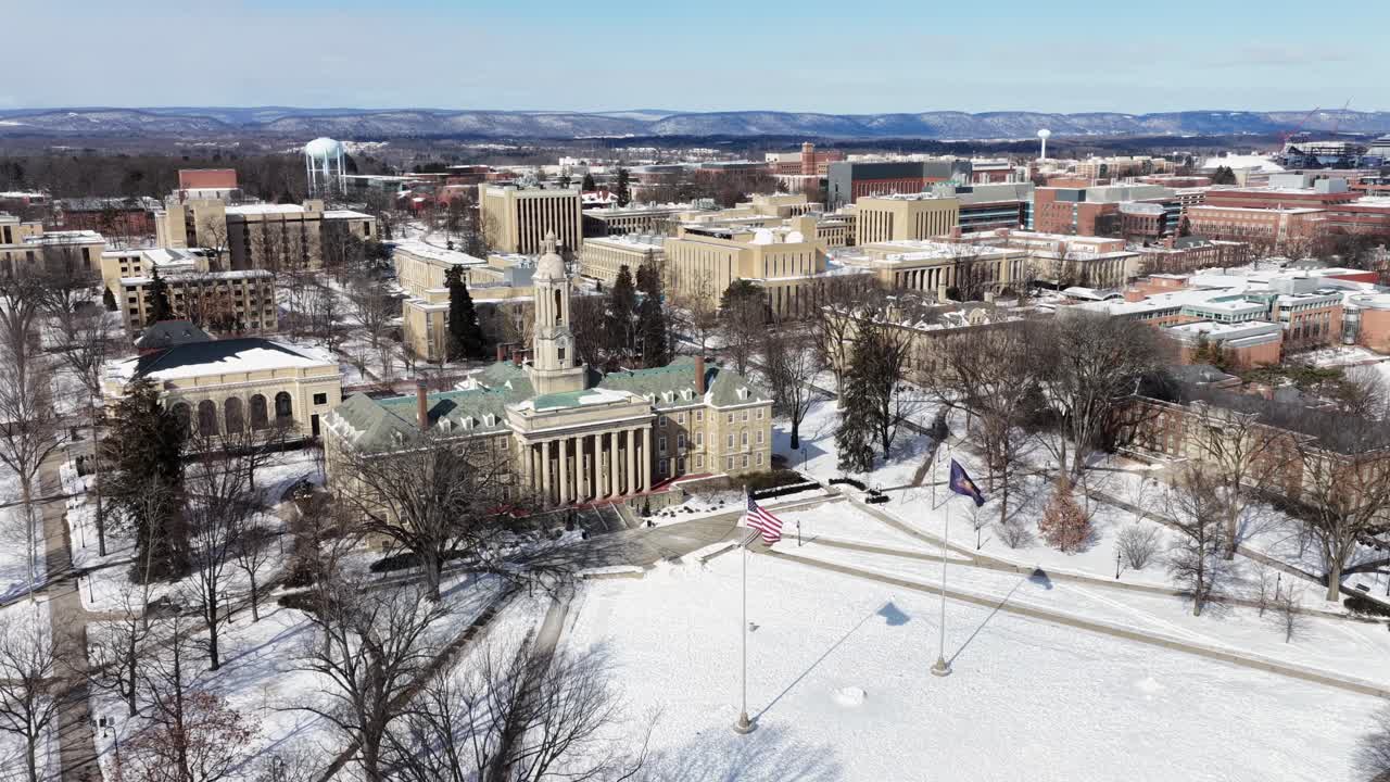Introducerende droneoptagelse af Pennsylvania State University på en snefyldt vinterdag. Viftende flag fra USA og PA på flagstang. Bredt luftfoto. Historiske bygninger på stor campus i universitetsby