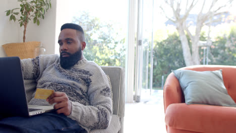 happy african american man using laptop and credit card in sunny living room, slow motion