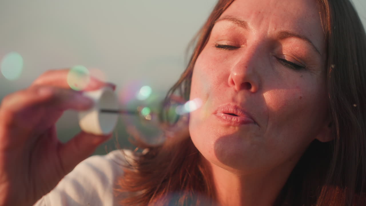 Close up of fair skinned woman blowing soapy bubble reflecting rainbow hues, gentle light illuminating lips and wand in soft focus outdoors, highlighting delicate bubble
