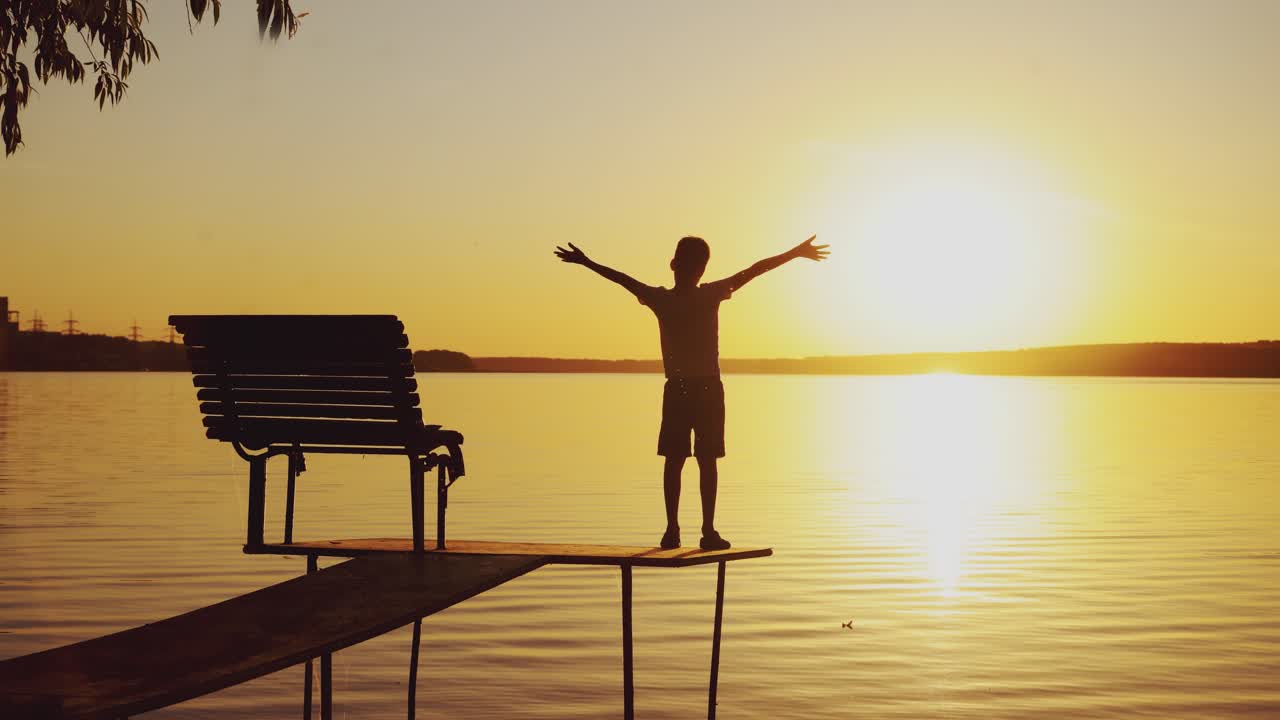 Boy doing yoga exercises by the river at sunset. Silhouette of a boy on an old bridge by the river. Beautiful summer sunset