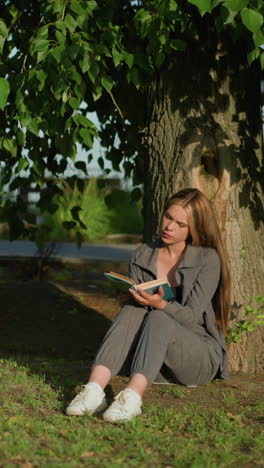 dama sentada al aire libre, cabeza ligeramente inclinada, apoyada en un árbol en un campo de hierba, leyendo un libro, las hojas de los árboles se balancean suavemente en la brisa, el fondo presenta vegetación y un riel de hierro ligeramente borroso