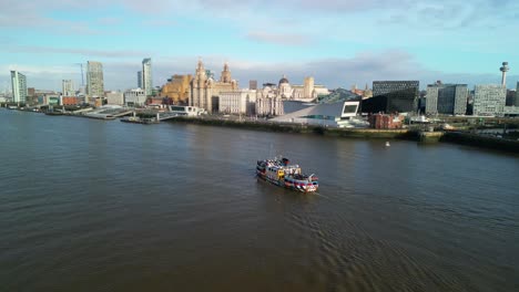 Ferry-crossing-the-River-Mersey,-Liverpool---approach-from-far,-tracked-by-drone-from-above-on-a-sunny-morning,-Liverpool-cityscape-backdrop