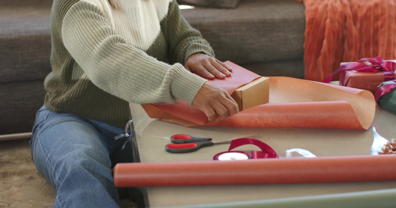 Woman placing box on orange paper, smoothing and folding at home table preparing Christmas gift