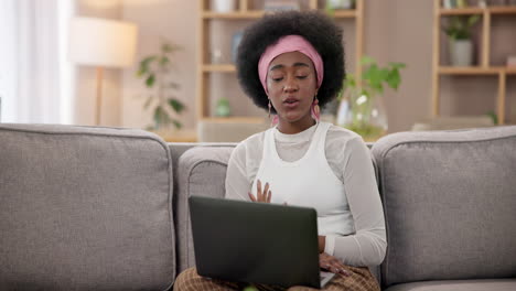 a young black woman sits on a couch in a living room, working on her laptop.