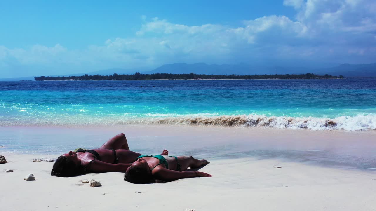 Two beautiful bikini girls lying in the sand and sunbathing on hot sunny day. Tropical vacation concept background