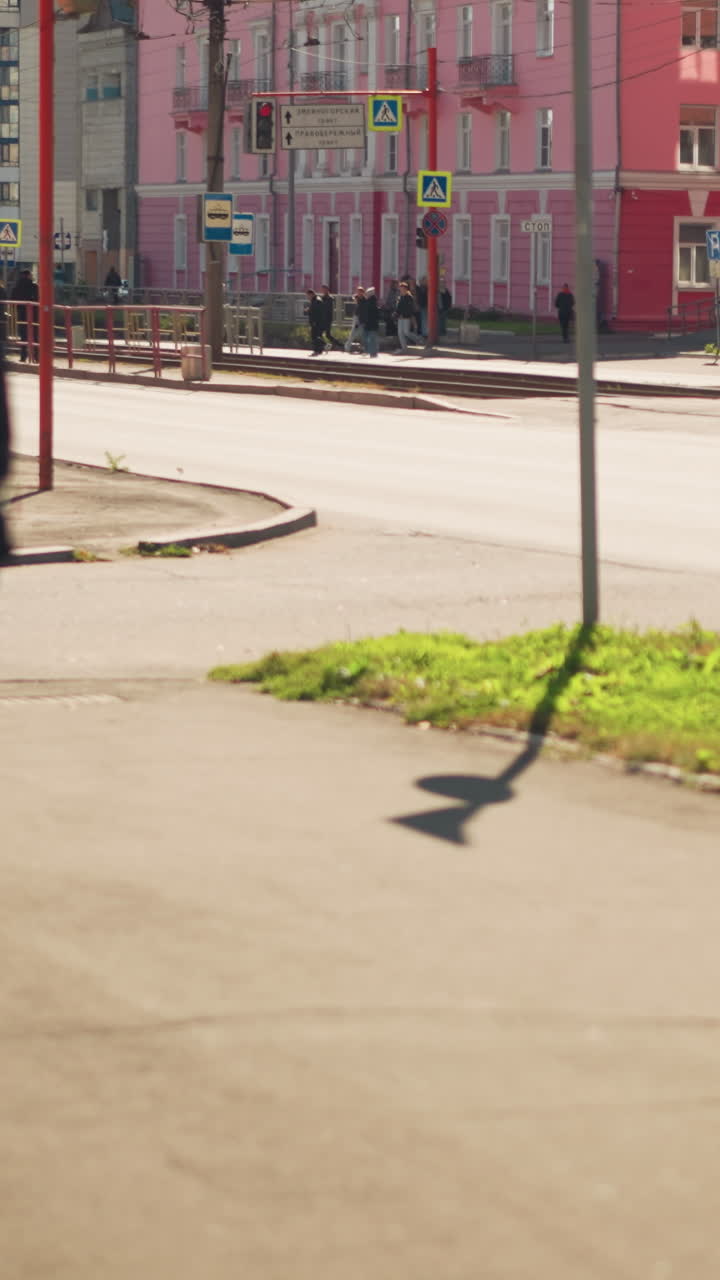 Hombre blanco corriendo por una intersección iluminada por el sol, pasando frente a escaparates rosas y señales de paso de peatones; un viajero apresurado mira su teléfono, la sombra se extiende por la acera, lleva una chaqueta informal y una mochila; ciudad de día