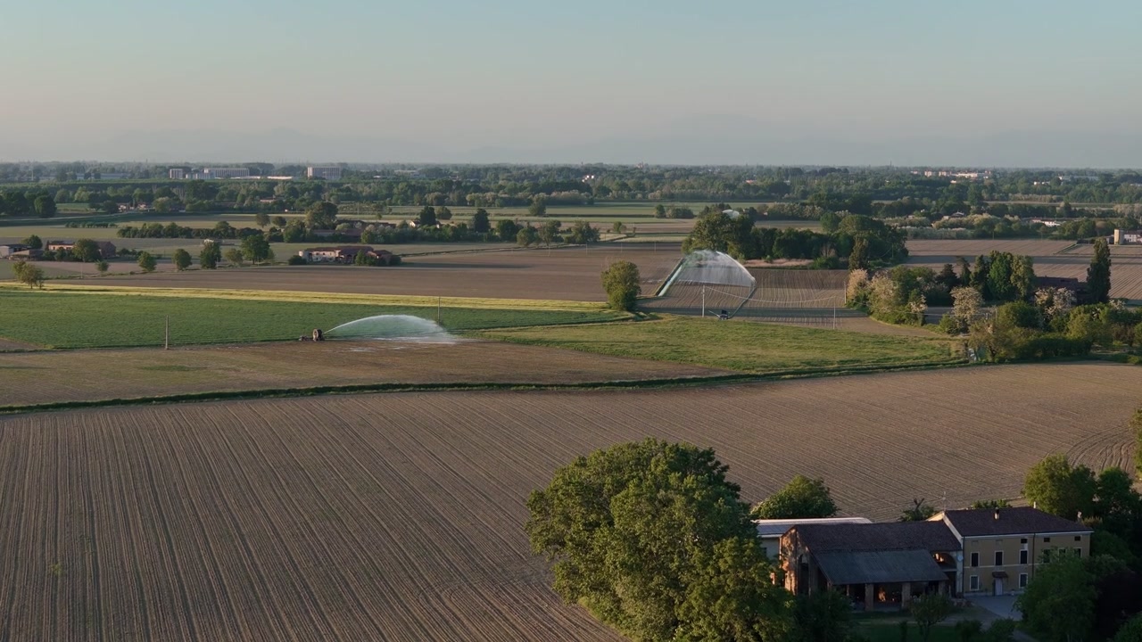 Traveling reel irrigators and sprinkler systems distribute water across spring farmland between Cremona Lombardy and Piacenza Emilia Romagna Italy near Po River floodplain, slow drone flyover