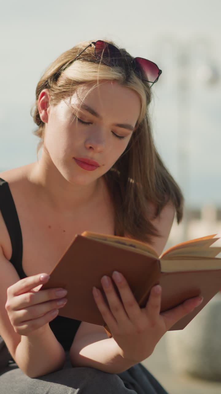 dama de solitario negro y gafas de sol descansando en su cabeza se sienta al aire libre, leyendo un libro cubierto de marrón pensativo como el viento levanta las páginas ligeramente, con un fondo borroso