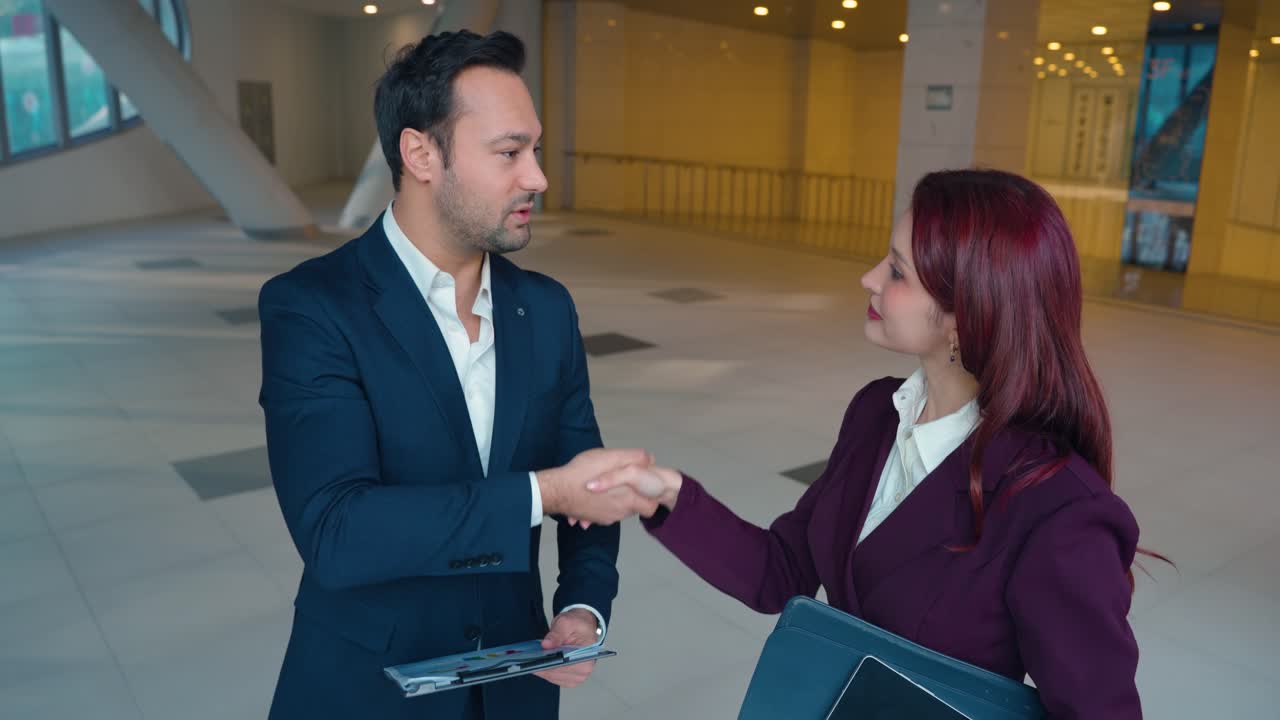 Empresario y empresaria se dan la mano cerrando un trato de negocios de pie en el moderno vestíbulo corporativo sosteniendo carpetas y un portapapeles