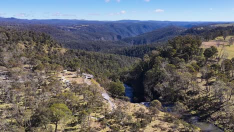drone glides above ebor gorge, revealing winding river, rugged terrain, and forested hills under clear daylight in new england, new south wales