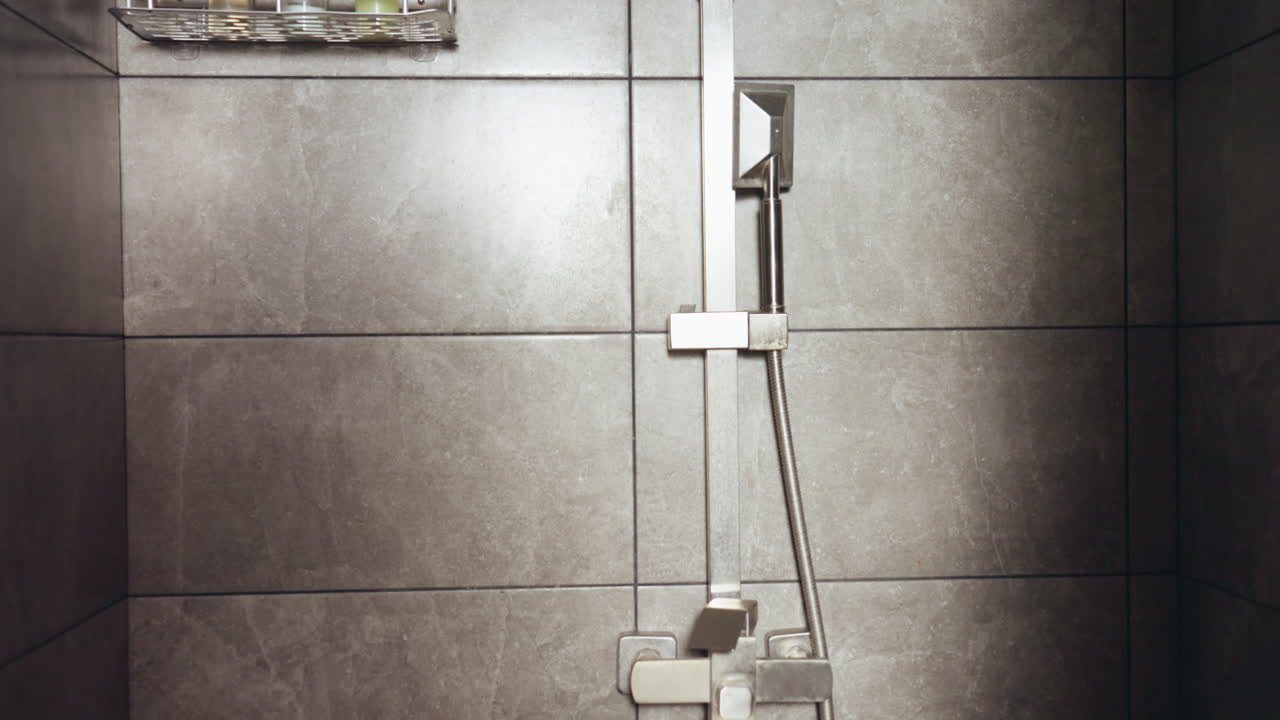 Modern tiled bathroom scene featuring ceiling-mounted square showerhead, wall mounted shelf with assorted cosmetic bottles, and compact ventilation fan, all set against clean gray ceramic tiles