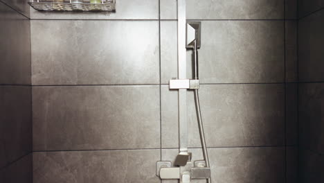 modern tiled bathroom scene featuring ceiling-mounted square showerhead, wall mounted shelf with assorted cosmetic bottles, and compact ventilation fan, all set against clean gray ceramic tiles