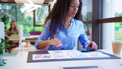 businesswoman sitting at desk in office taking photo of proofs or design layouts on mobile phone