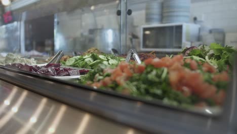 Vitrina-En-Una-Cafetería-Llena-De-Ensaladas-Recién-Hechas,-Con-Equipo-De-Cocina-Al-Fondo.