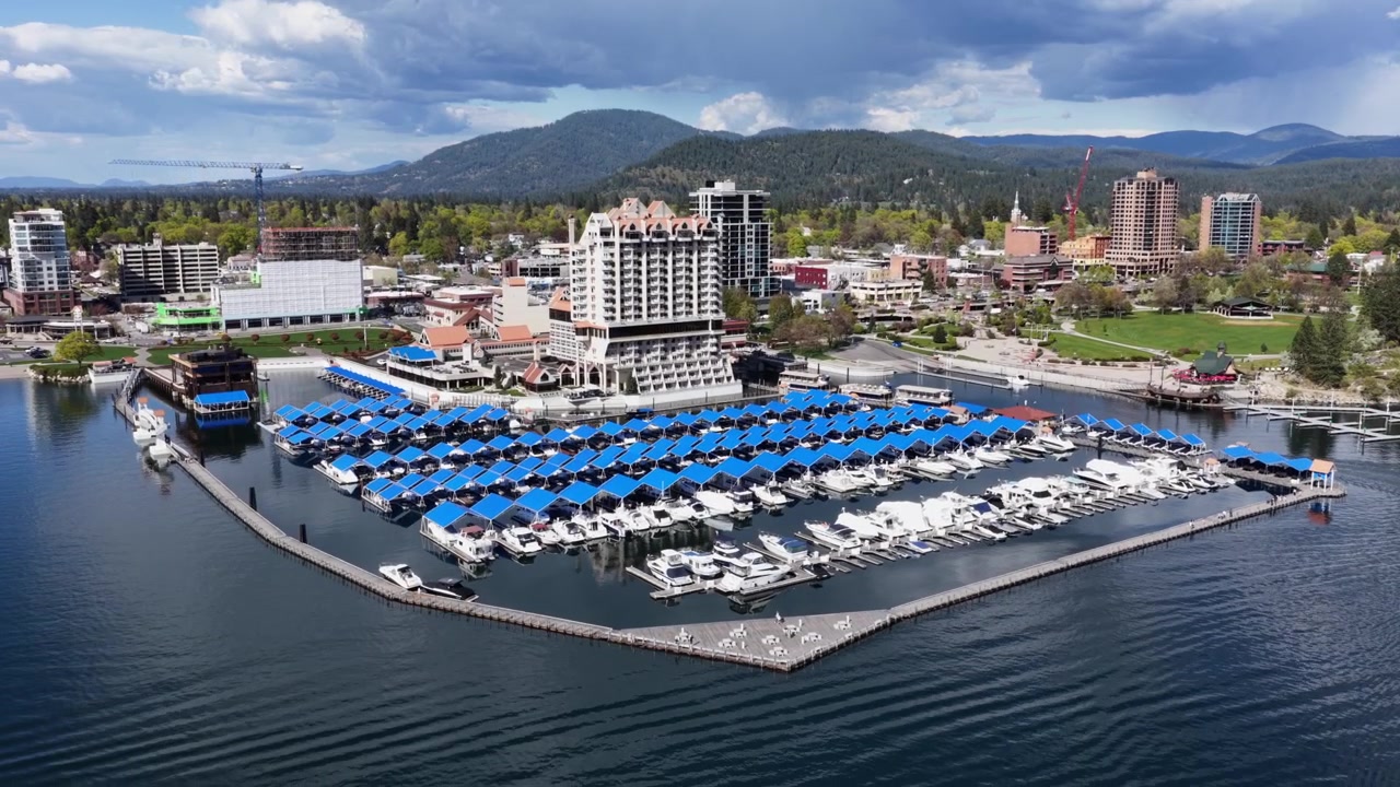 Lungolago di Coeur d'Alene con un grande porto turistico di lusso pieno di barche bianche sotto tettoie blu, la torre del Coeur d'Alene Resort, un parco verde e catene montuose di pini sotto un cielo estivo soleggiato. Ampia ripresa aerea orbitale.