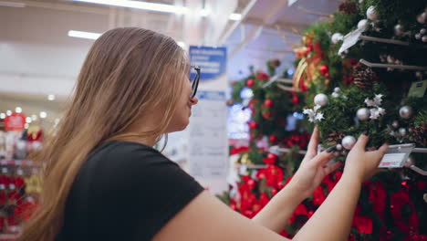 side view of woman with warm smile browsing holiday decorations in retail store, touching beautifully decorated wreaths with ornaments and pinecones, blurred shoppers in background