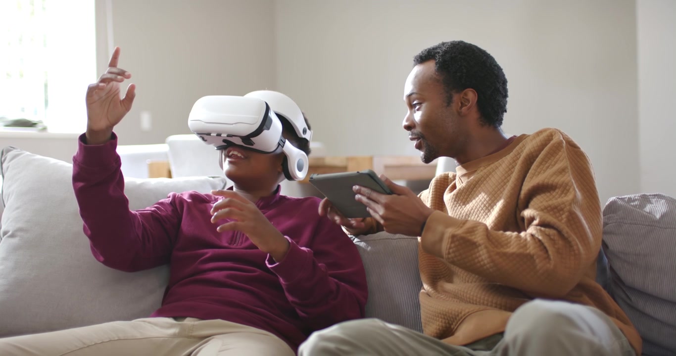 Padre afroamericano e figlio adolescente sul divano che consultano un tablet per guidare il figlio che usa un visore VR.