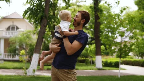 joven padre con barba en ropa casual está sosteniendo en los brazos a su pequeño hijo rubio. niño descalzo. hombre besando a su hijo. calle de cabaña en el fondo. verde y brillante
