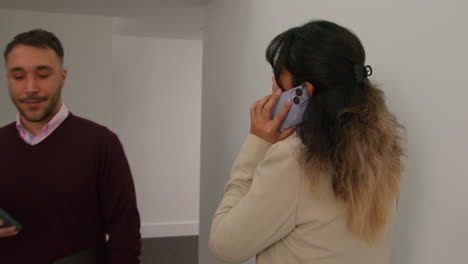 Female-Teacher-Talking-On-Mobile-Phone-Passing-Male-Colleague-As-They-Walk-Along-Corridor-In-School-Building