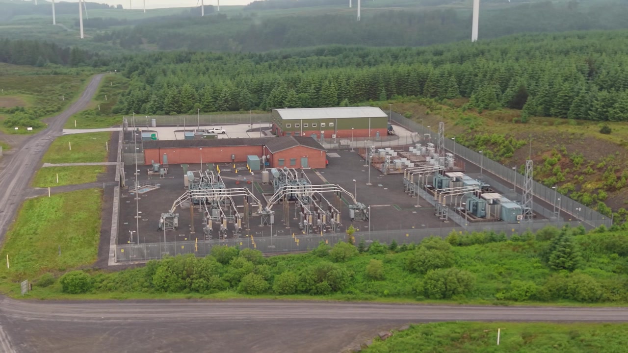 Elektrická rozvodna řídící a přesměrovávající tok elektřiny v přírodní krajině s větrnými turbínami v pozadí.