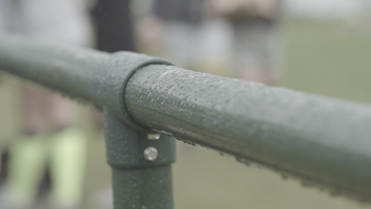 disparo de cerca de una barra o valla cubierta de gotas de lluvia con futbolistas en el fondo en registro de cámara lenta