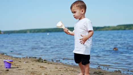lovely caucasian toddler in white t-shirt and black shorts on the beach. confused baby boy looks at his shovel and hand dirtied with sand.