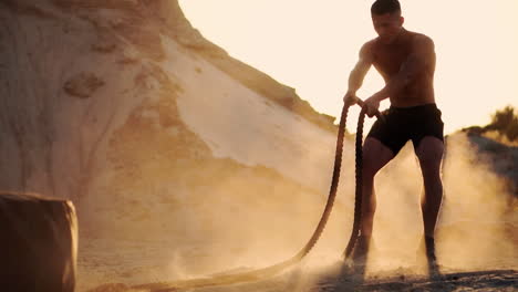 Atleta-Masculino-Haciendo-Flexiones-En-La-Playa-Y-Golpeando-La-Cuerda-En-El-Suelo,-Entrenando-Circularmente-Al-Sol-En-La-Playa-De-Arena-Levantando-Polvo-En-Cámara-Lenta.-Primer-Plano-De-Crossfit.