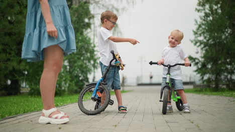 two happy siblings are seen pointing at each other while enjoying their time outdoors, they stand next to their bicycles, with a woman in a blue dress standing behind them