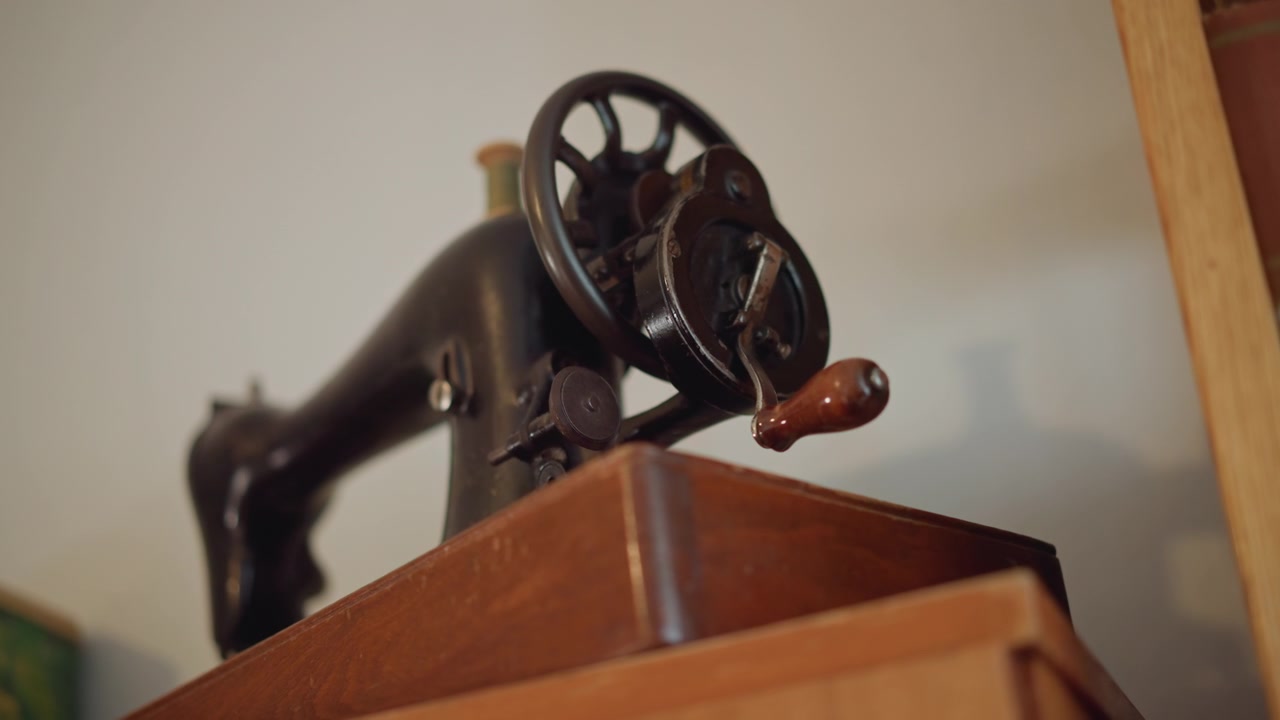 Antique sewing machine on wooden cabinet, visible hand crank and wheel, cast iron body with worn enamel, warm studio light, tailor workshop prop, heirloom decor, nostalgic craft mood