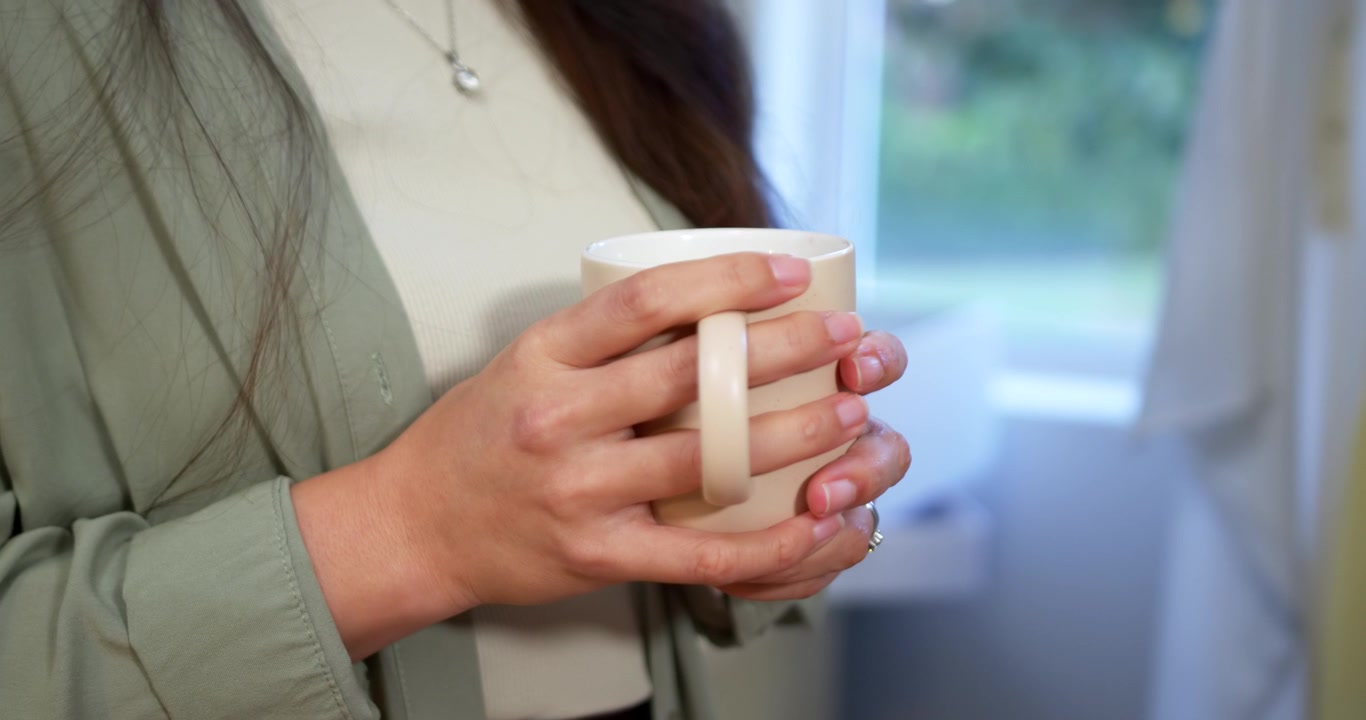 Mani che avvolgono una tazza di ceramica bianca, una donna la tiene vicino alla finestra mentre la stabilizza.
