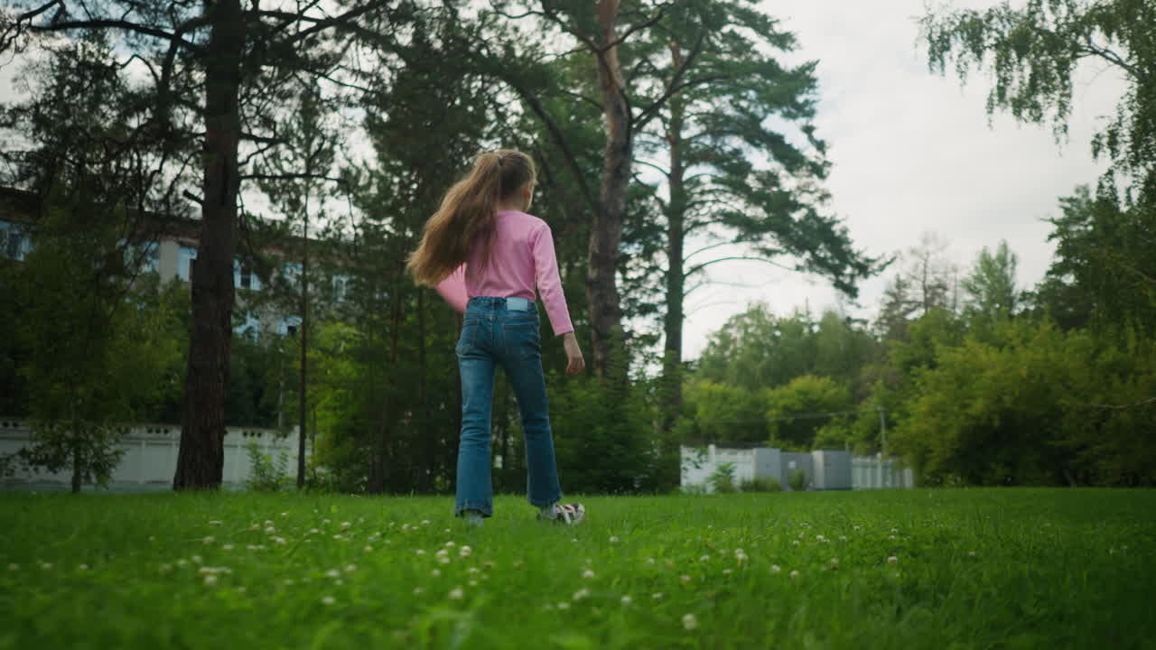 Back view of young girl in pink long-sleeve top and jeans walking across green grassy field while playfully swinging pink balloon, with apartment building and tall trees in background