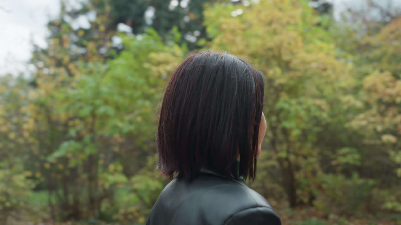 革のジャケットを着た女性が、緑のタートルネックを着て、ボブカットで物思いにふけりながら、秋の森を歩いている。落ち葉が敷き詰められた並木道、曇り空、自然なカラーパレット、静かで瞑想的な雰囲気、スローシネマティック。