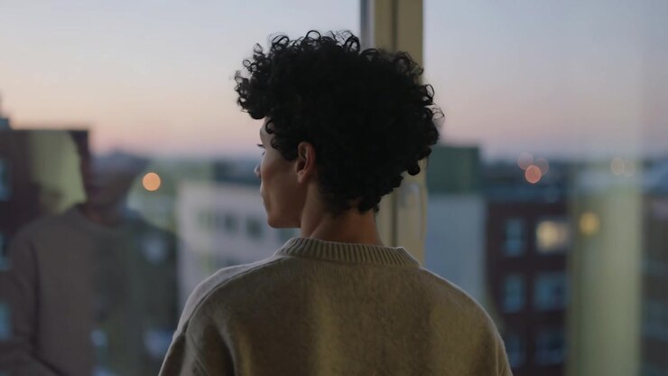 Dusk drawing man turning head and gazing out floor-to-ceiling window in high-rise, showing sweater