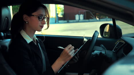 atractiva mujer de negocios caucásica en un coche estacionado, escribiendo notas en un clipboard, mientras se concentra y piensa