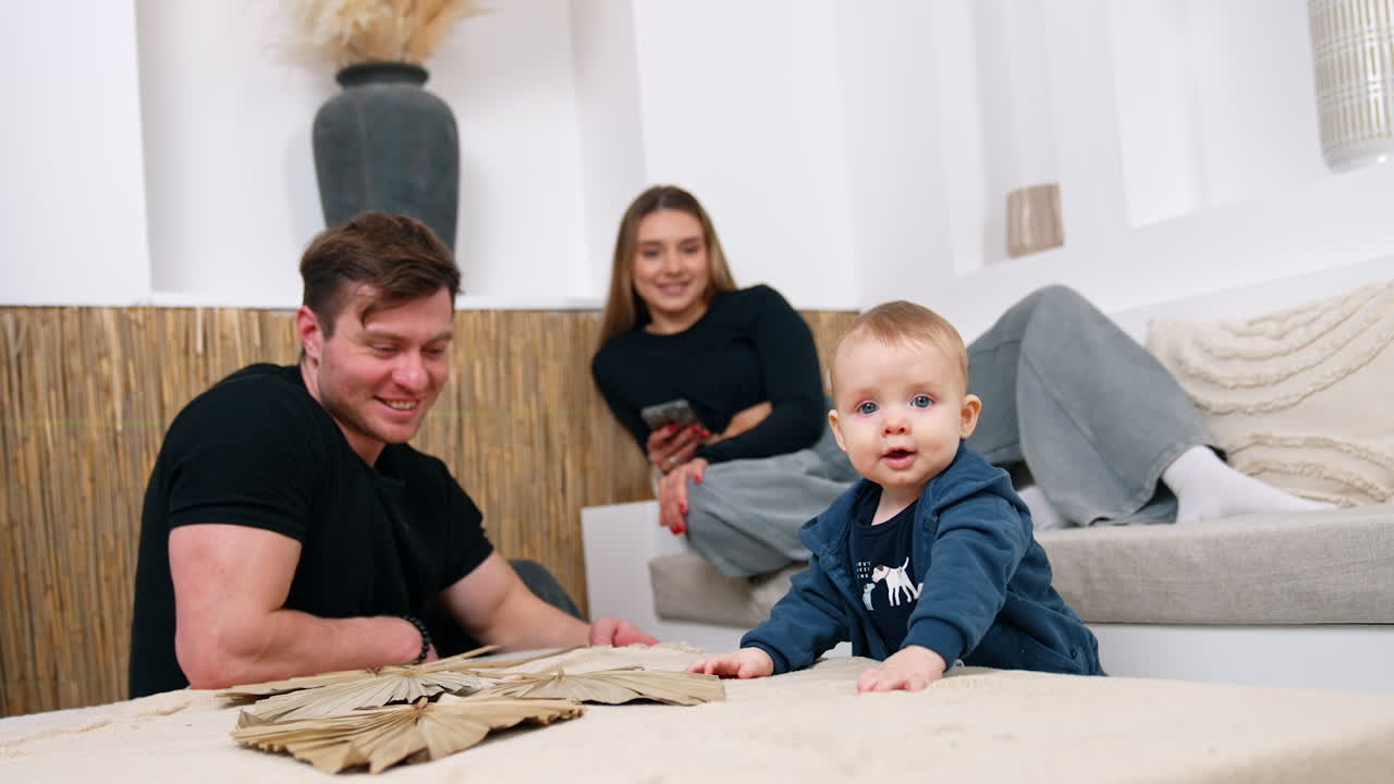 Sweet little baby boy in blue sweater stands at the coffee table. Smiling dad sits beside and mom sits on the sofa at backdrop.