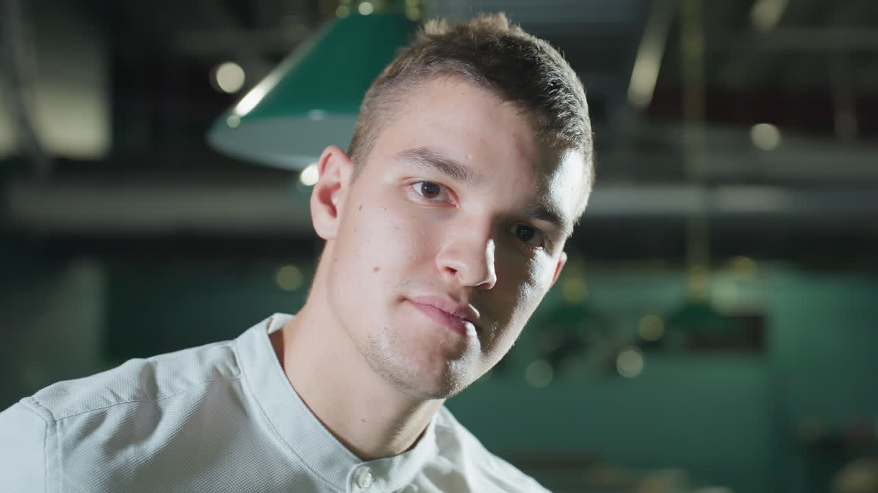 toma de primer plano de un hombre con camisa blanca con una ligera sonrisa en una sala de billar débilmente iluminada. las luces superiores crean reflejos suaves en su cara. la mesa de billar verde y las luces colgantes añaden profundidad a la escena